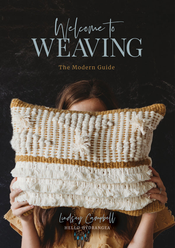 Person holding a textured, woven pillow in front of their face; the book cover reads Welcome to Weaving: The Modern Guide by Lindsey Campbell. The background is dark and the pillow features cream and mustard colors.