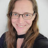 A woman with long brown hair and glasses smiles warmly at the camera in a black top against a light background, reflecting her passion for personal growth and the Guardian Tarot by Schiffer Publishing, Ltd.