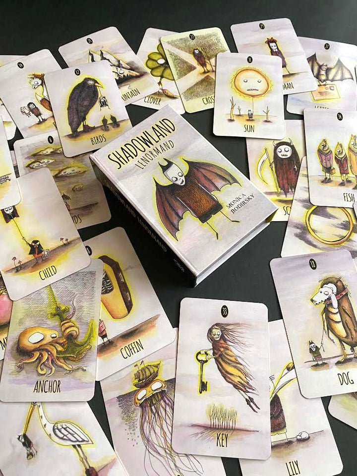 The Shadowland Lenormand by Schiffer Publishing, Ltd. features whimsical, surreal illustrations of a bat, key, sun, dog, fish, and owl—perfect for shadow work and cartomancy, shown spread on a black surface.