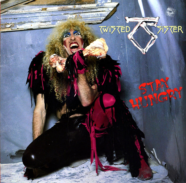 A person with long blond curly hair, heavy makeup, and a black and pink outfit holds a large bone in their mouth. Behind them, "Twisted Sister" and "Stay Hungry" are on a gray wall, as featured in The Art of Metal by Schiffer Publishing, Ltd.