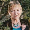 An older woman with short blonde hair stands outdoors among pine branches, smiling gently at the camera while holding the Steele Wizard Tarot by Schiffer Publishing, Ltd. She's dressed in a turquoise top, brown jacket, and statement turquoise necklace.