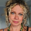 A woman with curly blonde-brown hair, blue eyes, and a nose ring wears a layered green beaded necklace and blue strap top. Schiffer Publishing’s Bonefire Tarot cards are visible in the blurred indoor background.