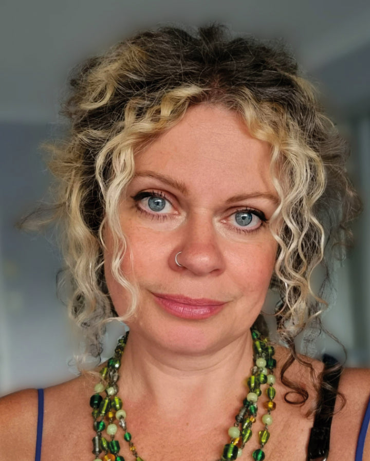 A woman with curly blonde-brown hair, blue eyes, and a nose ring wears a layered green beaded necklace and blue strap top. Schiffer Publishing’s Bonefire Tarot cards are visible in the blurred indoor background.