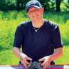 A person in a navy Seattle Seahawks cap and black shirt sits at a striped outdoor table, smiling as they shuffle Sigils tarot cards by Schiffer Publishing, Ltd., with greenery and yellow flowers in the background.