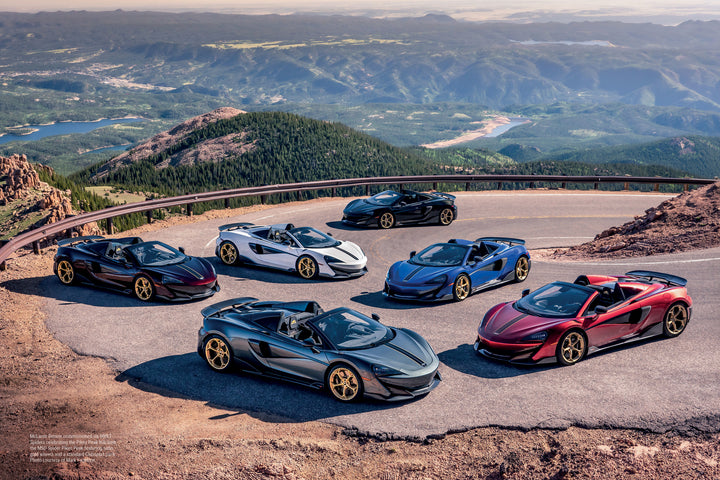 Six McLaren supercars by Schiffer Publishing, Ltd. are parked on a winding mountain road, overlooking a scenic valley with forests, lakes, and distant hills under a partly cloudy sky.