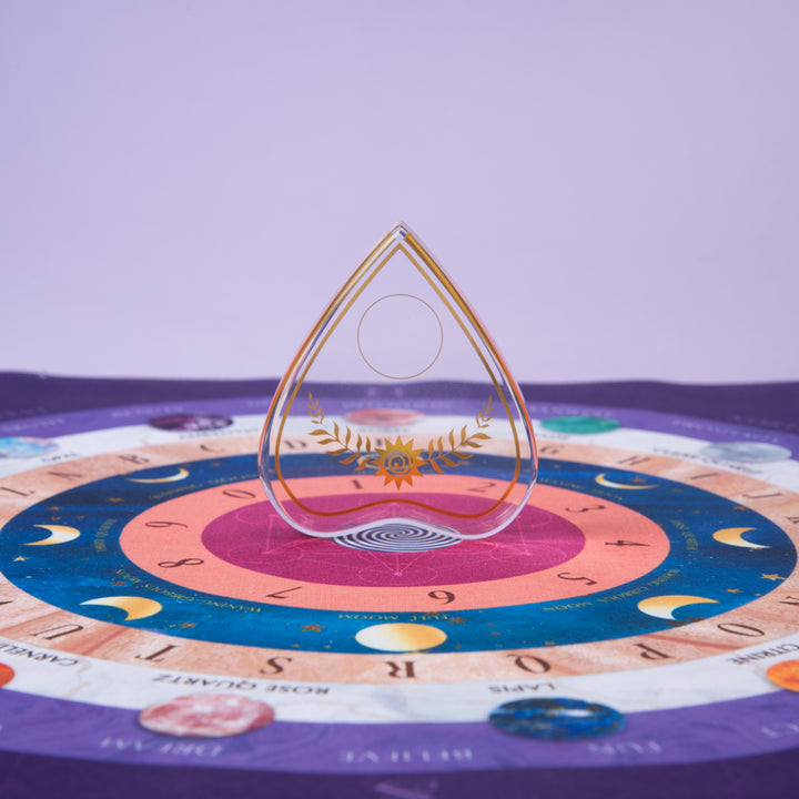 The Crystal Spirit Talking Board and Guidebook by Schiffer Publishing, Ltd. features a transparent gold-detailed planchette and a vibrant board with letters, numbers, and moon phases on a purple background inspired by sacred crystal energy.