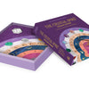 A partially opened purple box labeled "The Crystal Spirit Talking Board and Guidebook" by Schiffer Publishing, Ltd. reveals a vibrant spirit board adorned with gems, zodiac signs, numbers, and a clear quartz crystal for channeling crystal energy.