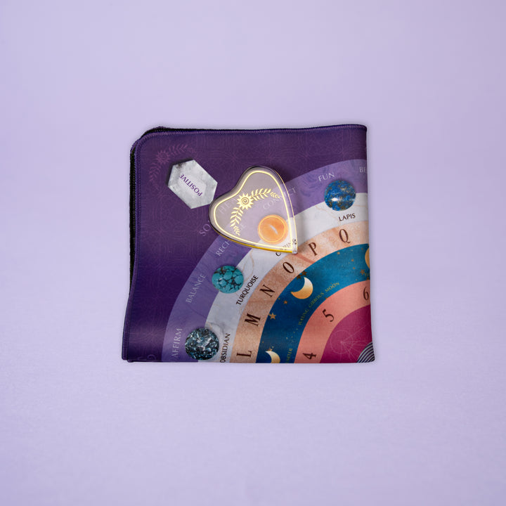 The Crystal Spirit Talking Board and Guidebook by Schiffer Publishing, Ltd. rests on a lavender surface, its colorful design featuring zodiac signs and gemstones; a gold heart-shaped sun-and-leaf pin sits on top.