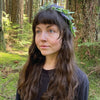 A woman with long brown hair and a leafy flower crown stands in a lush forest, reflecting the mystical spirit of The Animystic Tarot by Schiffer Publishing, Ltd.