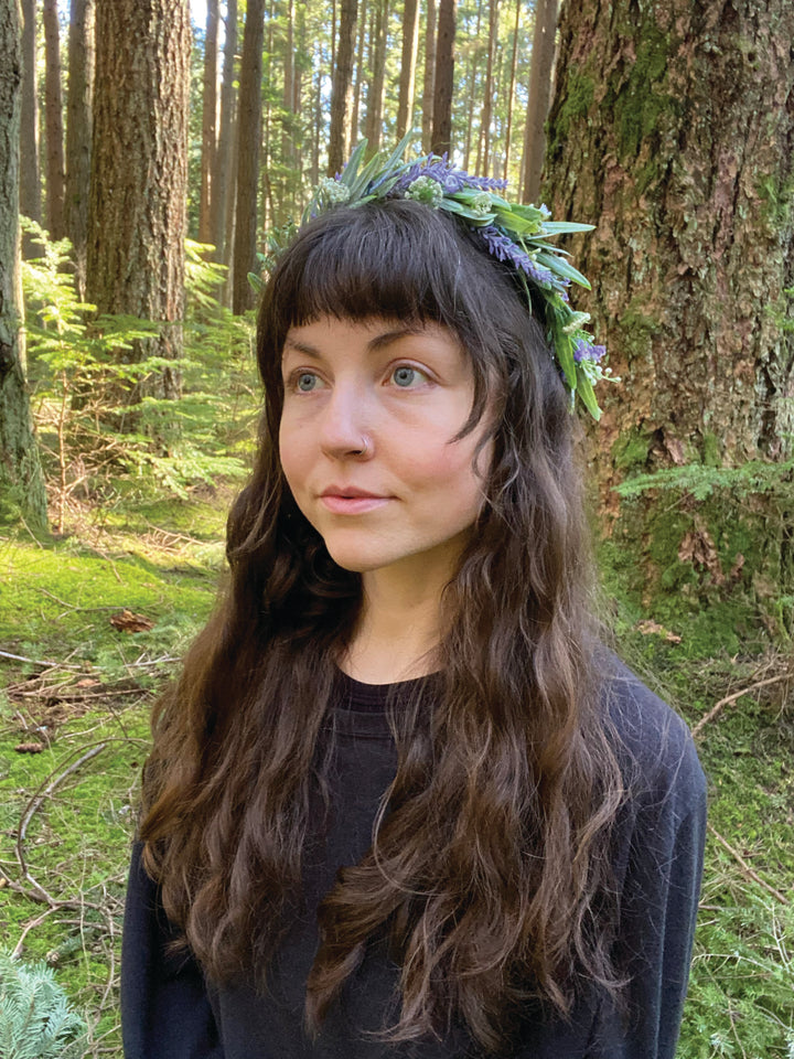 A woman with long brown hair and a leafy flower crown stands in a lush forest, reflecting the mystical spirit of The Animystic Tarot by Schiffer Publishing, Ltd.