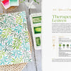 The Modern Watercolor Workshop by Schiffer Publishing, Ltd. lies open on a woven placemat, displaying botanical watercolor leaves with red berries—ideal for beginners. Nearby are paint, a green brush, candle, and towel; “Therapeutic Leaves” is shown on the right page.