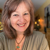A smiling woman with shoulder-length light brown hair wears a green top and a spiral pendant, standing indoors with warm lighting that reflects the earth-centered spirituality of the Gaian Tarot by Schiffer Publishing, Ltd.