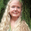 A woman with long, wavy blonde hair smiles softly before lush pines, evoking the serene beauty of a fine art tarot portrait from Boadicea's Tarot of Earthly Delights by Schiffer Publishing, Ltd.
