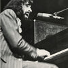 A bearded man with long hair passionately sings into a microphone while playing piano, evoking the legendary rock music captured in "Richard Manuel" by Schiffer Publishing, Ltd., as seen in this expressive black and white photo.