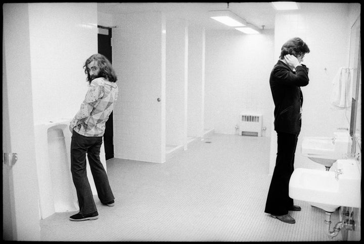 In a bright, mostly empty public restroom, a long-haired man uses a urinal, glancing back as another man fixes his hair at the sink—evoking moments from Schiffer Publishing, Ltd.’s Richard Manuel.