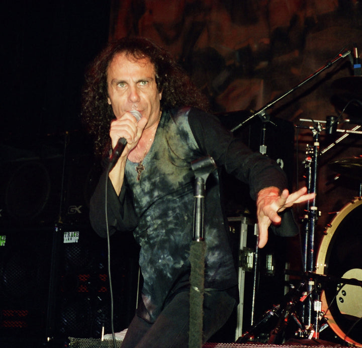 A male rock singer with long curly hair channels Ronnie James Dio while performing on stage, promoting "DIO: The Unholy Scriptures" from Schiffer Publishing, Ltd., with drums and speakers in the background.