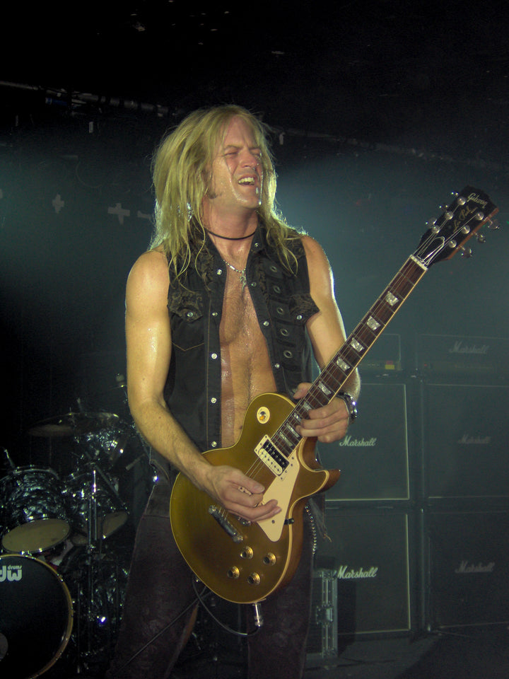A long-haired guitarist rocks on stage in a black vest, with drums and Marshall amps behind him—echoing the legendary Dio “Holy Diver” era, now captured in DIO: The Unholy Scriptures from Schiffer Publishing, Ltd.