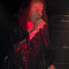 A male rock singer with long hair, channeling Ronnie James Dio of Dio, rocks a black shirt on stage with Marshall amps and lights behind him—capturing the spirit seen in "DIO: The Unholy Scriptures" from Schiffer Publishing, Ltd.