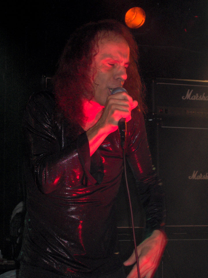 A male rock singer with long hair, channeling Ronnie James Dio of Dio, rocks a black shirt on stage with Marshall amps and lights behind him—capturing the spirit seen in "DIO: The Unholy Scriptures" from Schiffer Publishing, Ltd.