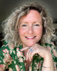 A smiling woman with curly blonde hair and a green floral shirt rests her chin on her hand, wearing a blue stone ring. She sits in soft natural light, ready to share "Yummy Watercolor" by Schiffer Publishing, Ltd. for beginners.