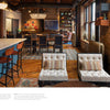 The "Midwest Home" by Schiffer Publishing, Ltd. showcases two tufted white lounge chairs, patterned pillows, a blue kitchen island with bar stools, exposed brick walls, large windows, unique art, and wood floors with decorative rugs.
