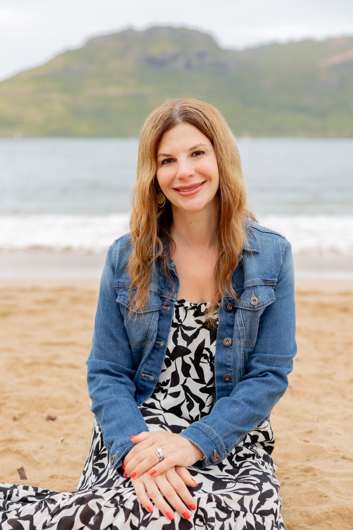 A woman with long, wavy hair sits on a sandy beach, smiling in a patterned dress and blue denim jacket. The ocean and green hills are dotted with shells from Seashells of Cape Cod and the Islands by Schiffer Publishing, Ltd.