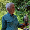 An older man in a green plaid shirt stands outdoors, holding The Peregrine Falcon by Schiffer Publishing, Ltd., with green trees in the background.
