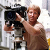 A man in a beige t-shirt and jeans films Memento Gorey interviews for Schiffer Publishing, Ltd. with a large camera on a tripod in an urban setting, tall buildings visible in the background.