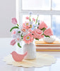 A white vase displays pink and white felt flowers made with the Bendable Blooms Chenille Stem Flower-Making Kit Cottage Garden by Schiffer Publishing, Ltd. Nearby is a pink bird ornament on a yellow placemat, with books and a dish by the window.