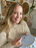 A smiling woman with long blonde hair holds the Sunny Stitches Embroidery Kit Sunny Days Ahead by Schiffer Publishing, Ltd., surrounded by embroidery art and supplies in a cozy, well-lit craft room.