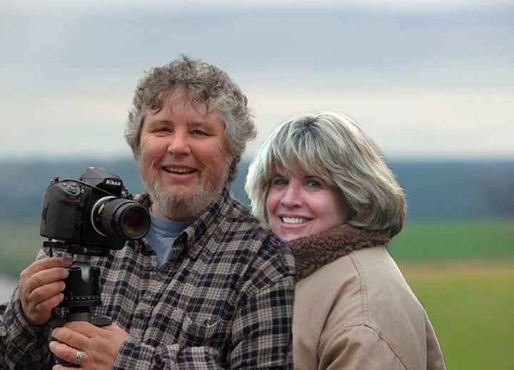 A man holding a camera on a tripod stands next to a smiling woman against the background of the Vineyards of Napa Valley book.