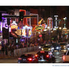 A busy Nashville travel souvenir street at night lined with bright neon signs for bars and music venues, with cars and taxis driving through the crowded Music City photography scene.