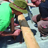 Two people work outdoors on a sustainable project inspired by "Natural Building" from Schiffer Publishing, Ltd., measuring a wooden log with pencil and string. One wears green; the other has sunglasses and dreadlocks. Others and tools are visible in the background.