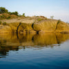 Rocky outcrops with sparse vegetation mirrored in calm blue water under a clear sky—a serene scene reminiscent of the photography in "Faces of the Susquehanna" from Schiffer Publishing, Ltd.