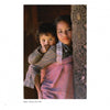 A young girl in “Face to Face” by Schiffer Publishing, Ltd. stands beside a carved post, carrying a child on her back with a pink cloth; both gaze gently at the camera in this striking portrait from Bhaktapur, Nepal, 1980.
