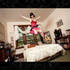 A person wearing a red corset and pink tutu jumps joyfully above a bed in a cozy bedroom, evoking burlesque vibes—capturing the playful spirit of "It's All That Glitters" by Schiffer Publishing, Ltd., among bookshelves and large windows.