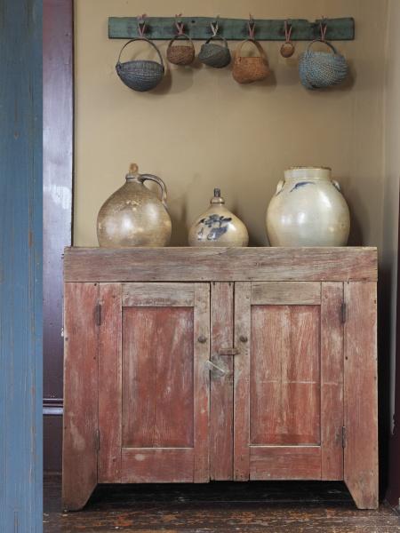 In "Passion for Primitives" by Schiffer Publishing, Ltd., a rustic red wooden cabinet with ceramic jugs and a rack of woven baskets captures the inviting charm of primitive décor in a cozy, vintage setting.