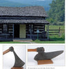 A grassy field with hills frames a log cabin and wooden porch. Below, Everyday Artifacts by Schiffer Publishing, Ltd. features close-up photos of a vintage broadax axe—an early American artifact—shown from two angles.