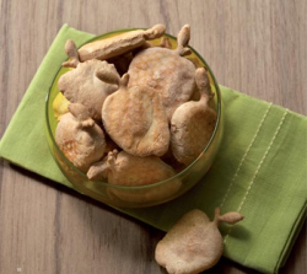 A glass bowl of pear-shaped dog treats sits on a green cloth atop wood, with one treat beside it—a creative recipe idea from "Cooking for Dogs" by Schiffer Publishing, Ltd.