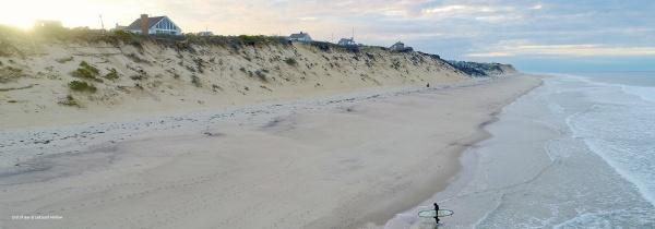 A drone photo from "Cape Cod and the Islands" by Schiffer Publishing, Ltd. captures a sandy beach with gentle waves, a surfer carrying a board by the shore, and houses on a grassy bluff under a partly cloudy sky.