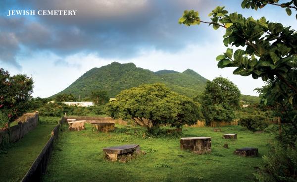 A lush cemetery with stone gravestones, trees, and mountains under a cloudy sky. "Jewish Treasures of the Caribbean" by Schiffer Publishing, Ltd. explores one of the oldest Jewish communities.