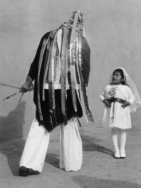 A scene from "Pueblo Dancing" by Schiffer Publishing, Ltd. shows a person in traditional Pueblo attire with ribbons facing a young girl in a white dress and veil, evoking the depth of Native American history.