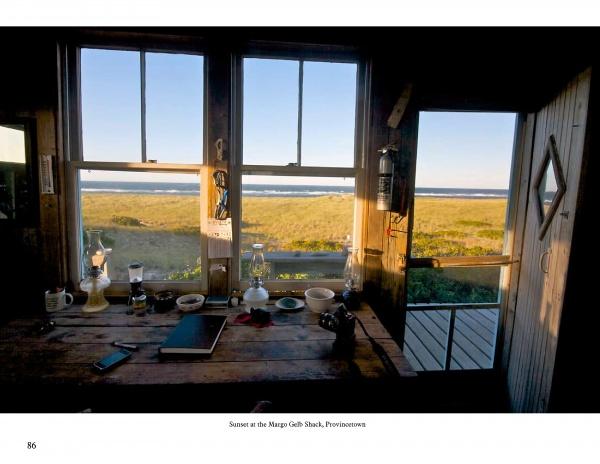 Cape Cod & the Islands Reflections by Schiffer Publishing, Ltd. captures classic Cape Cod scenes: a rustic desk with books and lamps, large windows framing grassy dunes and ocean views, all bathed in sunset light at the Margo Gath Shack, Provincetown.