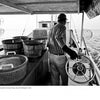 A man steers a boat amid baskets and barrels in this black and white image from "Chesapeake Views" by Schiffer Publishing, Ltd., evoking the daily life and fishing traditions of the Chesapeake Bay.