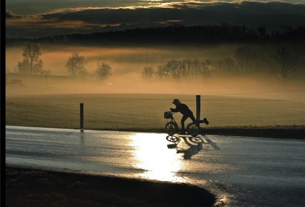 A cyclist rides a wet road at sunrise in Lancaster County, their reflection visible as mist drifts over fields—capturing the peaceful Amish life depicted in Faces of Lancaster County Postcards from Schiffer Publishing, Ltd.
