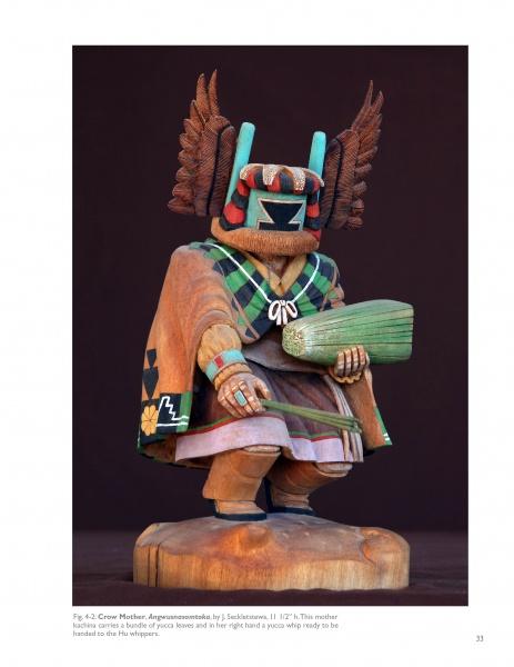 The Hopi Kachinas by Schiffer Publishing, Ltd. features a wooden doll adorned with a detailed mask, vibrant attire, feathers, and accessories like a corn cob and rattle, standing on a wooden base against a dark background.