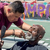 A barber trims an elderly man's beard outdoors as he relaxes in a black cape. Behind them, colorful graffiti and a basketball court embody the spirit of Design with Love by Schiffer Publishing, Ltd., highlighting vibrant community development.