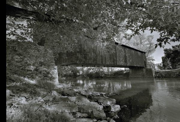 A black-and-white photo featured on the Bridges of Lancaster County Postcards by Schiffer Publishing, Ltd. shows a covered bridge over a calm river, framed by rocks and trees with reflections, capturing timeless postcard beauty.