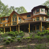 The Cedar Style by Schiffer Publishing, Ltd. features a spacious two-story log cabin with peaked roofs and a wraparound porch, nestled among trees on a landscaped hillside—highlighting timeless log home charm.