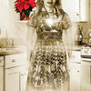 A sepia-toned image of a smiling woman in a floral dress stands in a vintage kitchen, a glowing heart on her chest. Poinsettias and greenery decorate the scene from A Christmas Tarot by Schiffer Publishing, Ltd.